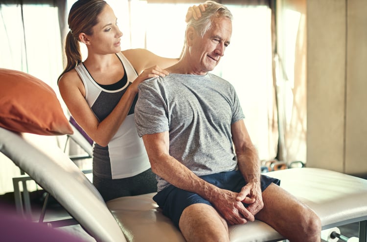 Senior man sitting on massage table with masseuse wondering what holistic health care is