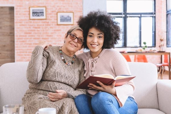 two women sitting on a couch reading a book and smiling for a photo